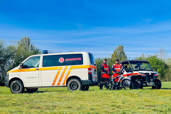 Ein Johanniter-Mannschaftstransportwagen, ein UTV-Geländefahrzeug, zwei Johanniter und ein Mountain-Bike auf einer grünen Wiese