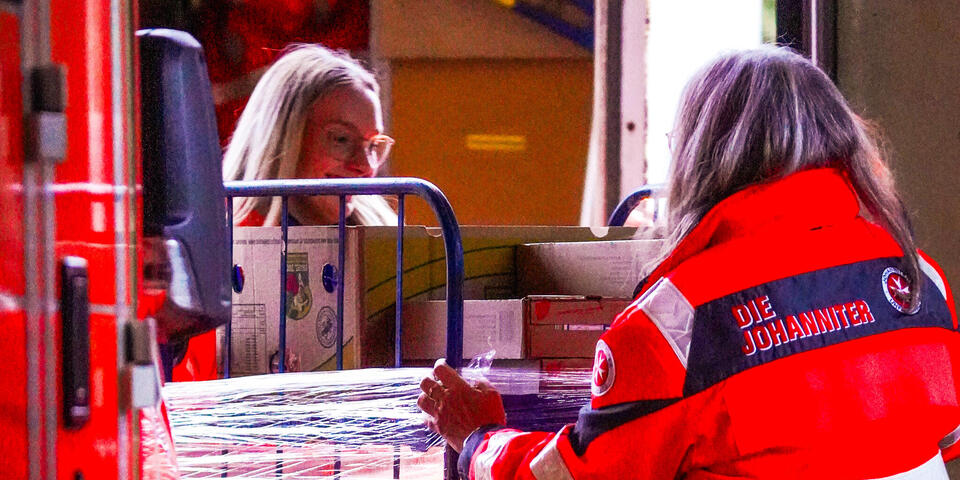 Zwei Frauen in Johanniter-Jacken schieben einen mit Waren gefüllten Rollbehälter