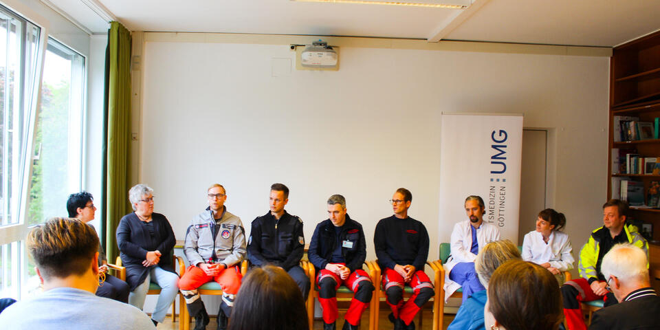 Zwei Frauen, mehrere Männer in Rettungsdienstbekleidung und ein Mann und eine Frau in Arztkitteln sitzen in einer Reihe vor Publikum.
