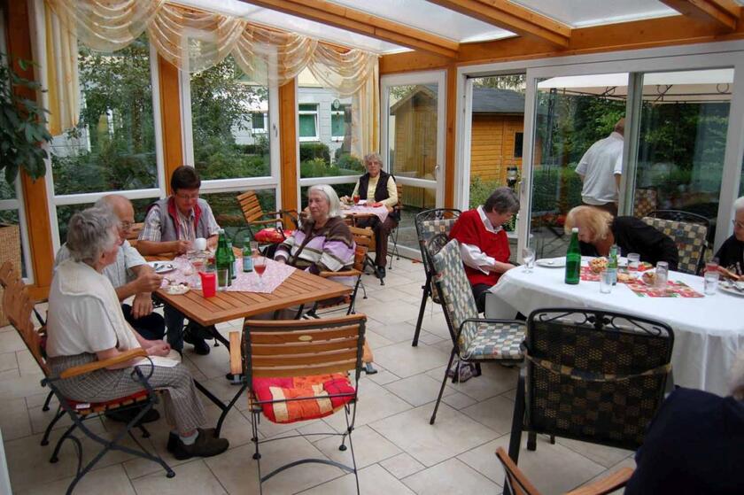 Die Senioren sitzen an Tischen im lichtdurchfluteten Wintergarten der Tagespflege und unterhalten sich.
