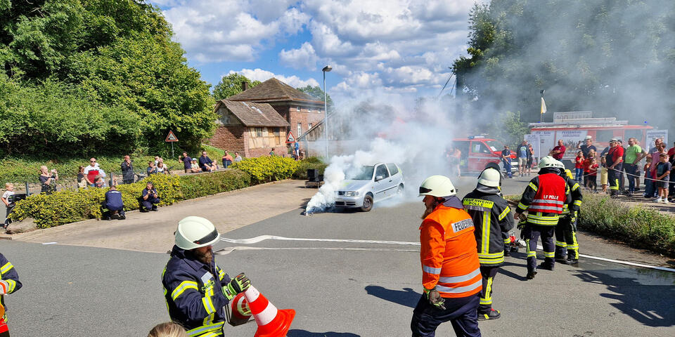 Eine Vorführung: Feuerwehr- und Polizeikräfte. Im Hintergrund ein vermeintlich brennender Wagenr