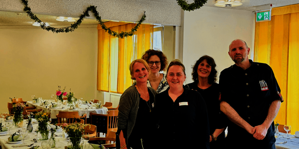 vier Frauen und ein Mann stehen lächelnd neben einem festlich gedeckten Tisch mit Blumen. An den Dekcen hängen grüne Girlanden, die Gardinen sind sonnengelb.