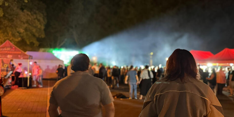 Ein Johanniter und eine Johanniterin schauen auf eine Menschenmenge. Es ist Nacht und im Hintergrund ist eine Bühne mit Lichteffekten