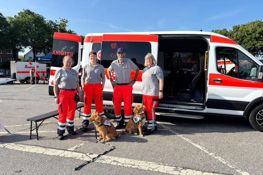 Ehrenamtliche posieren mit zwei Retungshunden vor einem Johanniter Einsatzfahrzeug auf dem Heider Marktplatz.