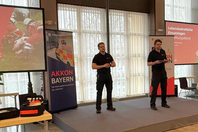 Zwei Personen stehen auf einer kleinen Bühne und berichten über Lebensbedrohliche Einsatzlagen im Rettungsdienst. Im Hintergrund sind Beamer und Aufsteller zu sehen, die Präsentationen und Bilder zeigen.  