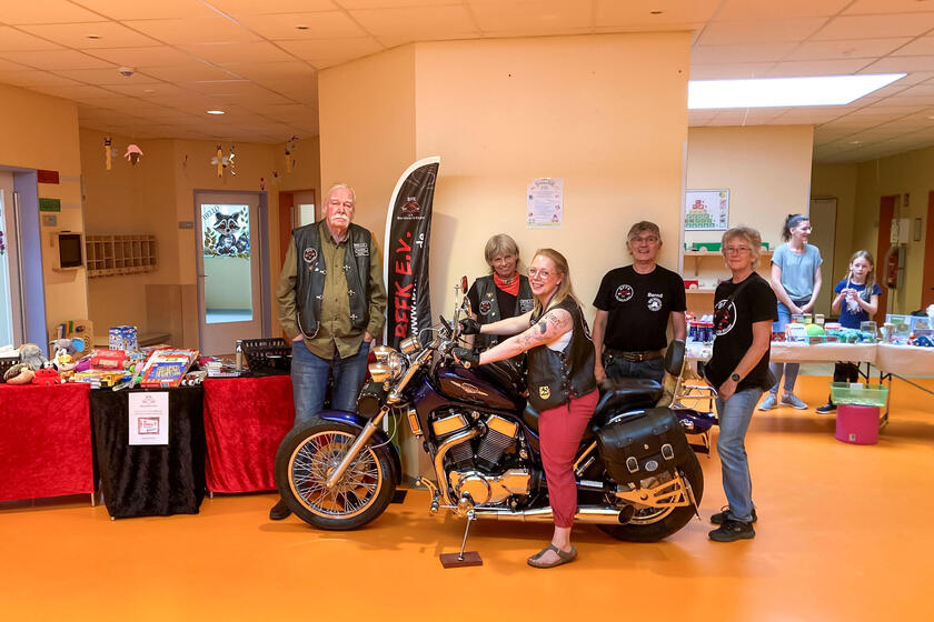 Mitglieder des Biker-Vereins stehen neben dem Stand. Eine Mutter sitzt auf einem Motorrad.