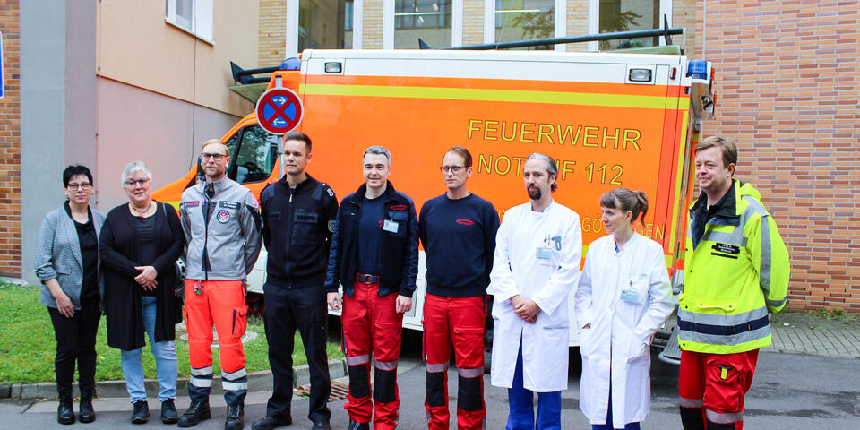 Zwei Frauen, mehrere Männer in Rettungsdienstbekleidung und ein Mann und eine Frau in Arztkitteln stehen vor einem Rettungswagen.