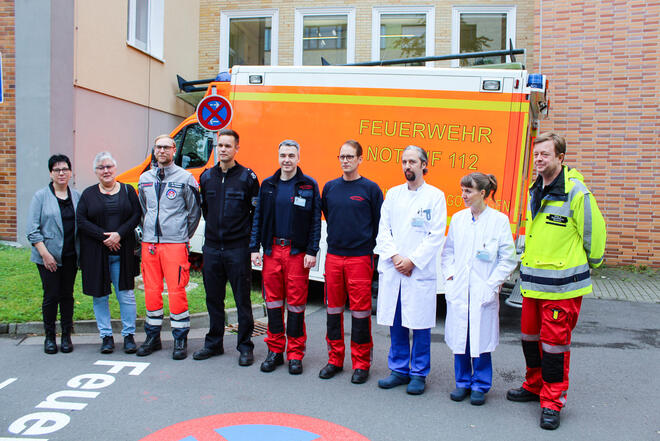Zwei Frauen, mehrere Männer in Rettungsdienstbekleidung und ein Mann und eine Frau in Arztkitteln stehen vor einem Rettungswagen.
