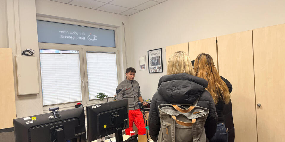 Zwei Schüler und eine Schülerin hören einem jungen Mann in Johanniter-Bekleidung zu