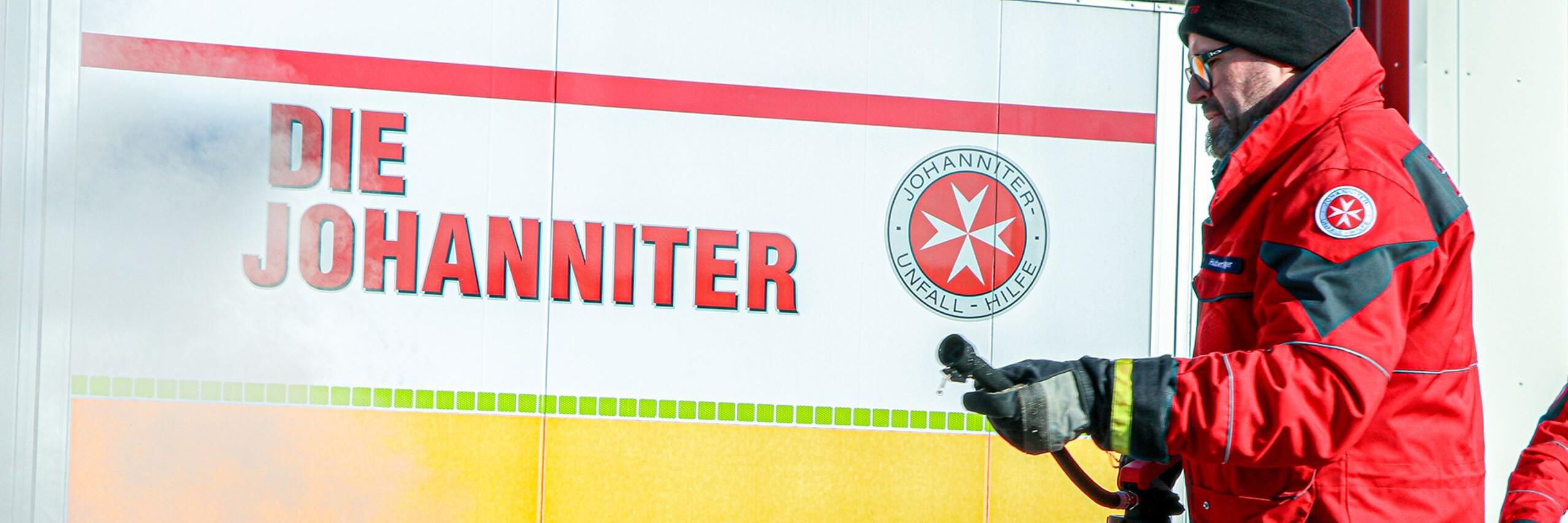 Johanniter in roter Einsatzkleidung löscht mit Feuerlöscher ein Übungsfeuer vor Fahrzeug mit Aufschrift „Die Johanniter“.