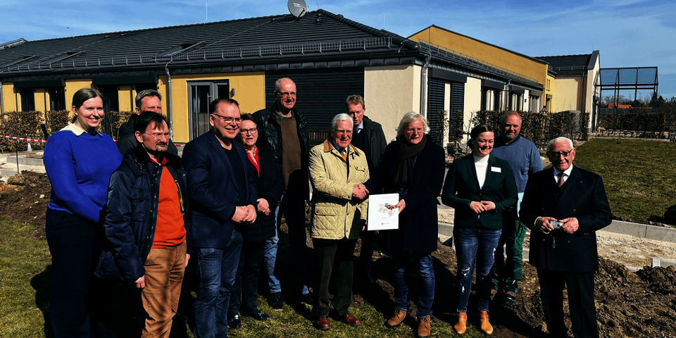 Zwölf Menschen stehen auf einer Grasfläche vor einem flachen Gebäude. Zwei halten ein Blatt Papier und reichen sich die Hände.
