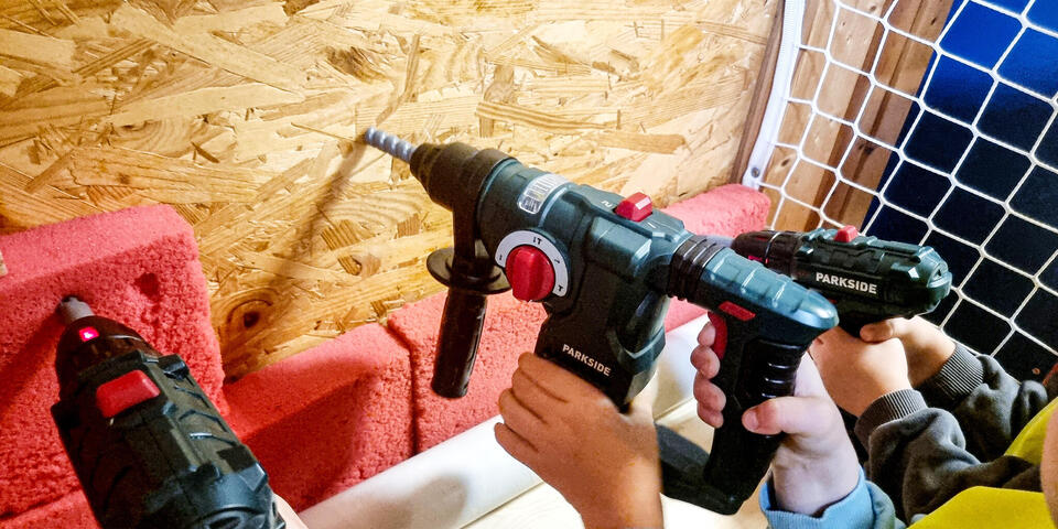 Kinderhände mit Spielzeugbohrern an einer Wand aus Spanplatten
