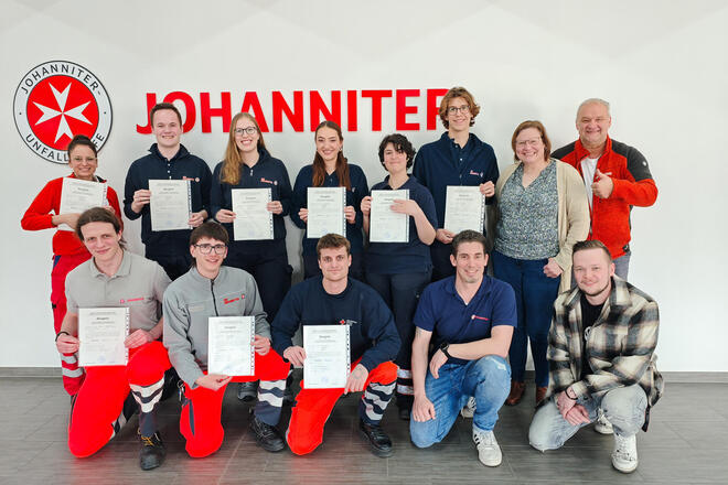 Vor einer Wand mit Johanniter-Logo stehen acht Personen, sechs davon halten eine Urkunde in der Hand. Vor ihnen knien fünf Personen, drei davon halten eine Urkunde in der Hand.