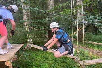Ein Kletterer überquert hockend ein an Seilen hängendes Brett.