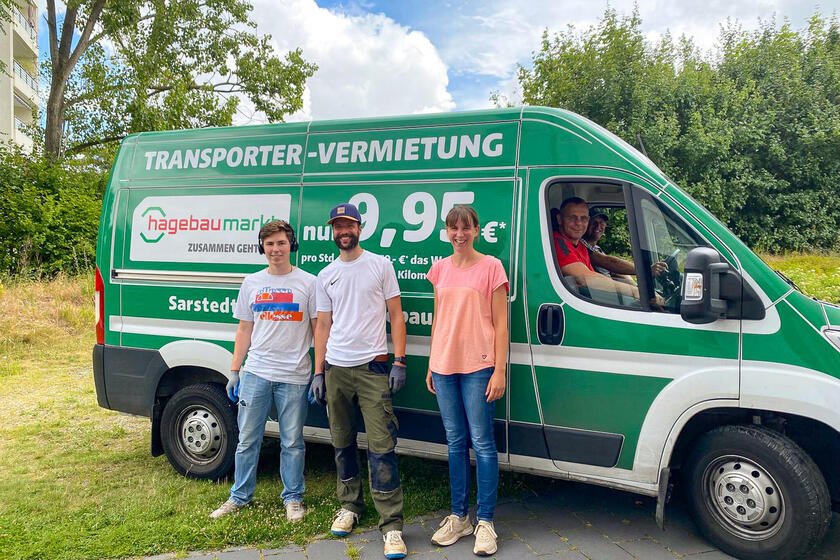 Zwei Männer und eine Frau vor einem grünen Transporter auf dem "Hagebaumarkt" steht. Im Transporter sitzen zwei weitere Männer.