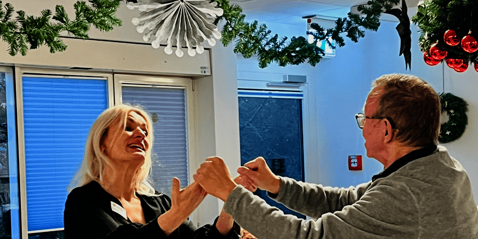 Eine Frau und ein Mann tanzen einander anlächelnd in einem weihnachtlich geschmückten Raum, im Hintergrund weitere Menschen am Tisch.