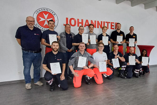 Neun Männer und drei Frauen in Rettungsdienstbekleidung mit einer Urkunden. Ein Mann und eine Frau mit Johanniter-Bekleidung stehen daneben. Im Hintergrund ein großes Johanniter-Logo. 