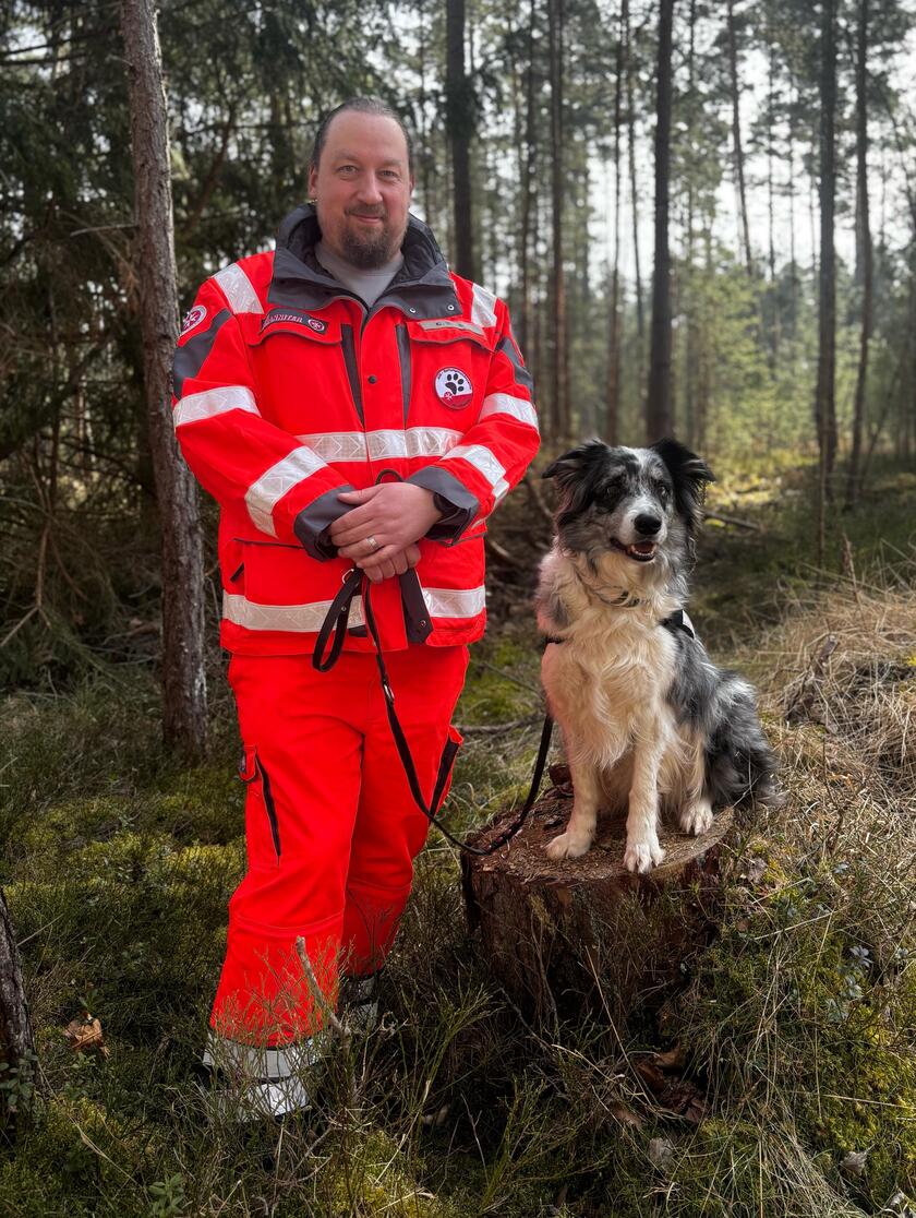 Ein Johanniter-Rettungshundeführer steht in leuchtender Einsatzkleidung im Wald. Neben ihm sitzt sein schwarz-weißer Rettungshund aufmerksam auf einem Baumstumpf. Beide blicken freundlich in die Kamera.