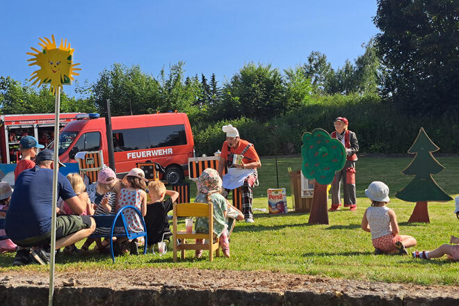 Kinder auf einer Wiese, die sich den Auftritt von Opa Krause ansehen, im Hintergrund steht die Freiwillige Feuerwehr und es scheint die Sonne bei strahlend blauem Himmel. 