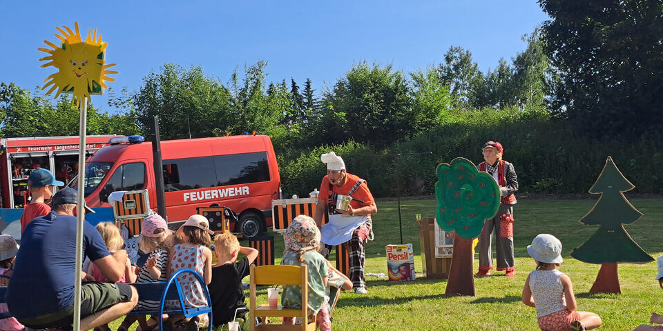Kinder auf einer Wiese, die sich den Auftritt von Opa Krause ansehen, im Hintergrund steht die Freiwillige Feuerwehr und es scheint die Sonne bei strahlend blauem Himmel. 