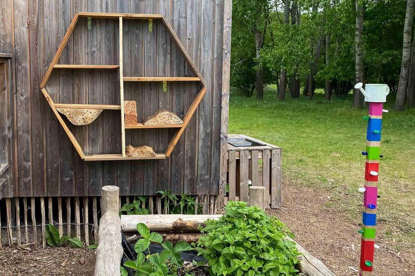 Ein noch unfertiges Insektenhotel hängt unter dem Schild der Kita an einer Wand. Davor steht ein Hochbeet.