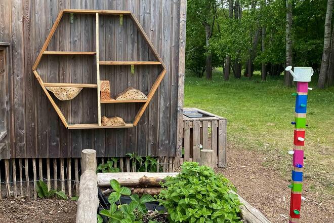 Ein noch unfertiges Insektenhotel hängt unter dem Schild der Kita an einer Wand. Davor steht ein Hochbeet.