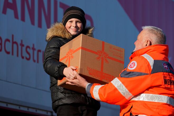 Ein Johanniter in Einsatzkleidung überreicht einem ca. 10-jährigen Jungen einen großen Karton - das Johanniter-Weihnachtstrucker-Paket. Im Hintergrund steht ein großer LKW mit dem Schriftzug Johanniter-Weihnachtstrucker.