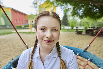 Eine junge Frau sitzt auf einer Netzschaukel und lächelt in die Kamera. Im Hintergrund sind ein Spielplatz und Bäume zu erkennen, die eine ruhige und freundliche Atmosphäre vermitteln.