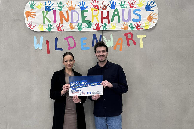 Charlotte Pieslinger De Bernardin übergibt einen Spendenscheck an Einrichtungsleiter Johannes kern vor einer Grauen Wand mit einem Schild der Einrichtung umrahmt von bunten Handabdrücken.