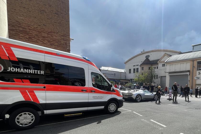 Ein Rettungswagen der Johanniter steht am Straßenrand in einer Stadt. Im Hintergrund sind mehrere Menschen auf einer Straße sowie Häuser zu sehen. Ein paar Menschen begutachten Sportwägen.