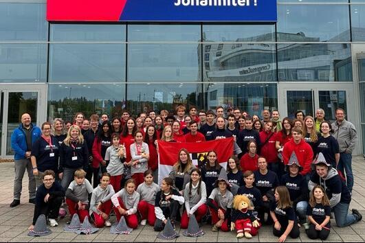 Gruppe von Johanniter-Mitgliedern, bestehend aus Kindern, Jugendlichen und Erwachsenen, posiert lächelnd vor der Messe in Erfurt. Im Hintergrund ist auf einem großen digitalen Display das Johanniter-Logo zu sehen, dazu die Botschaft: „Wer sind die besten Rettungskräfte im ganzen Land? – Na klar, wir Johanniter!“. In der ersten Reihe halten mehrere Teilnehmende eine rot-weiße Flagge, vorne sitzt ein Maskottchen in Johanniter-Kleidung.