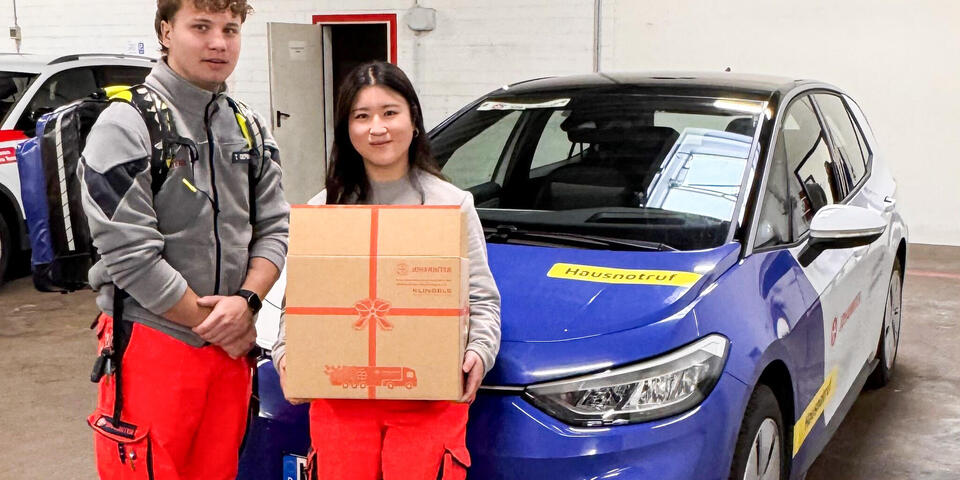 Ein junger Mann und eine junge Frau in Einsatzkleidung vor einem Johanniter-Auto. Sie hält ein Paket in der Hand.