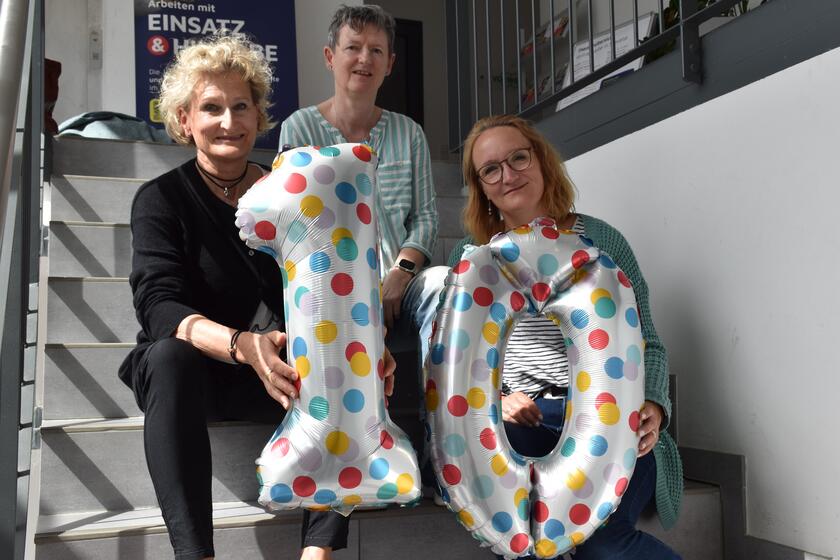 drei Mitarbeiterinnen des Besuchsdienst sitzen auf der Treppe und halten Luftballons in Form einer Zehn hoch