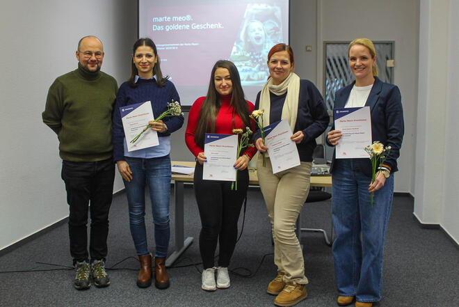 Vier Frauen mit einem Zertifikat in der Hand und ein Mann in einem großen Raum. Im Hintergrund eine Präsentation auf einer Leinwand. 