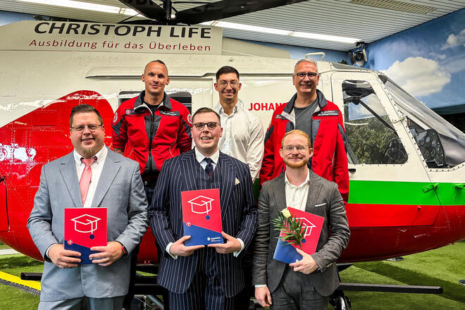 Im Vordergrund drei Männer mit einer Urkunde. Dahinter drei Männer, zwei davon mit einer Johanniter-Jacke. Im Hintergrund ein Helikopter in einer Halle. 