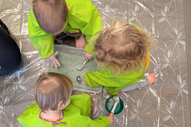 Kinder sitzen auf einer Plane. Sie haben grüne Kittel an und spielen mit einer grünen Masse. 