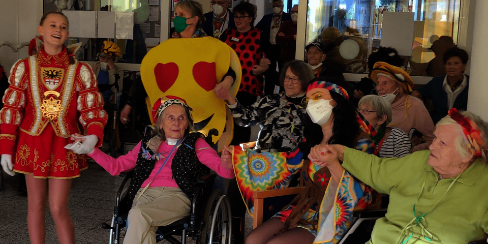 Ein rotgekleidetes Tanzmariechen hält die Händen der neben ihr im Sitzen schunkelnden Senioren