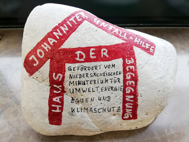 Auf dem weißen Stein ist ein rotes Haus mit dem Text "Haus der Begegnung" aufgemalt. Auf dem Dach des Hauses steht "Johanniter-Unfall-Hilfe".