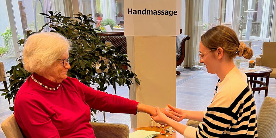 Zwei Frauen sitzen gegenüber an einem kleinen Tisch. Die jüngere Frau hält die Hand der älteren in den Händen.