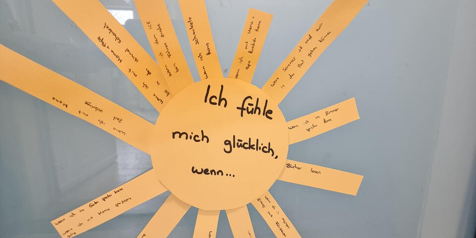 Eine Sonne aus Pappe auf der steht "Was mich glücklich macht". Auf ihren Strahlen stehen Beispiele geschrieben. 