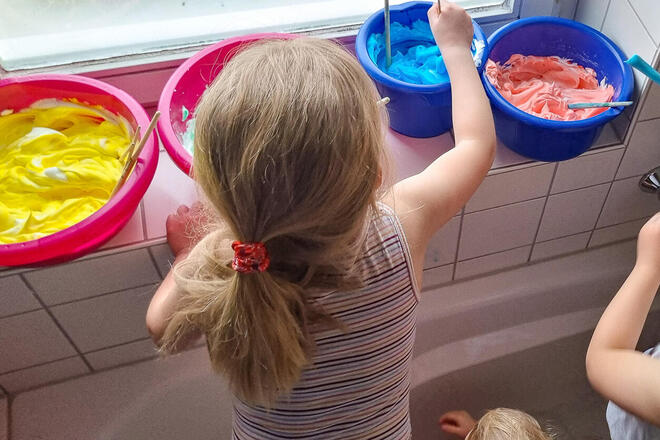 Ein kleines Mädchen rührt in einer Wanne stehend Farben in bunten Plastikschalen an.