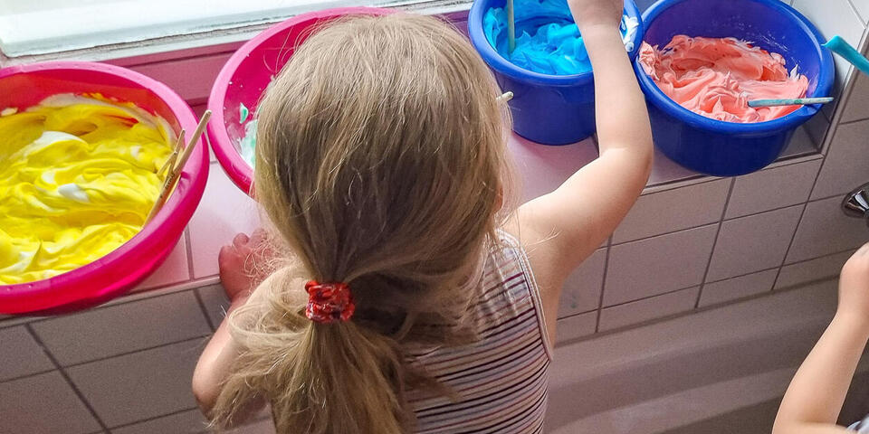 Ein kleines Mädchen rührt in einer Wanne stehend Farben in bunten Plastikschalen an.