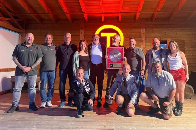 Zwölf Menschen versammeln sich in einem Holzhaus. In ihrer Mitte präsentieren sie eine große Auszeichnung mit einer silbernen Schallplatte. Im Hintergrund leuchtet ein rundes Symbol an der Wand als T abgebildet. Die Atmosphäre ist festlich.