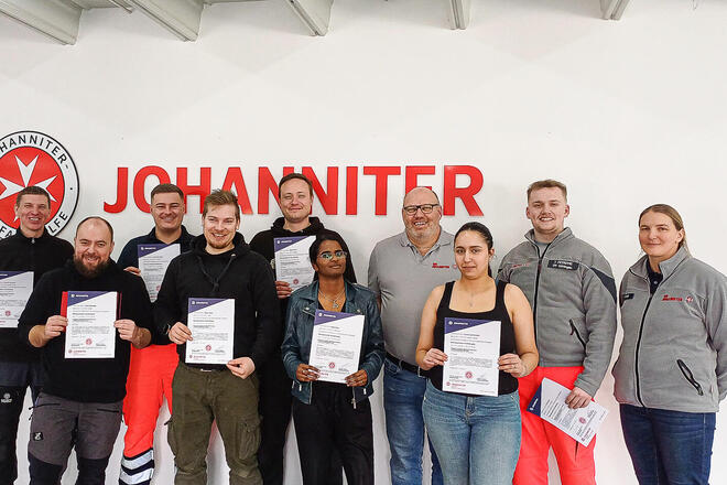 Sechs Männer und zwei Frauen mit Zertifikaten in der Hand. Daneben ein Mann und eine Frau in Johanniter-Bekleidung. Alle stehen vor einer Wand mit dem Schriftzug und Logo der Johanniter 