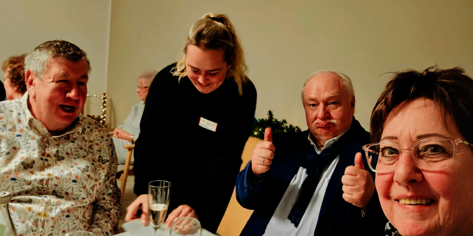 Gäste an einem festlich gedeckten Tisch, eine Servicekraft serviert, ein Gast zeigt Daumen nach oben, fröhliche Stimmung.