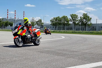 Zwei Einsatzkräfte der Johanniter-Motorradstaffel üben mit ihren Einsatzmotorrädern auf einem Trainingsplatz bei sonnigem Wetter. Sie tragen gelbe Helme und reflektierende Kleidung.