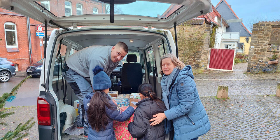 Kinder und eine Frau laden Päckchen in ein Johanniter-Auto. Ein Johanniter steht im Fahrzeug und hilft.