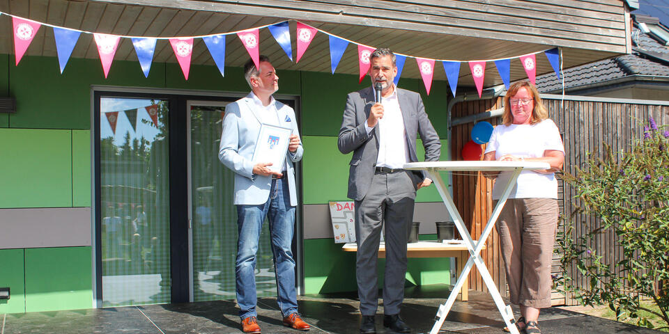 Ein Mann mit Anzug und Mikro neben ihm ein Mann und eine Frau