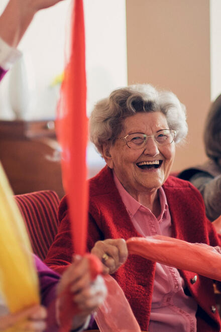 Lachende auf dem Stuhl sitzende Seniorinnen bewegen sich spielend mit einem bunten Tuch