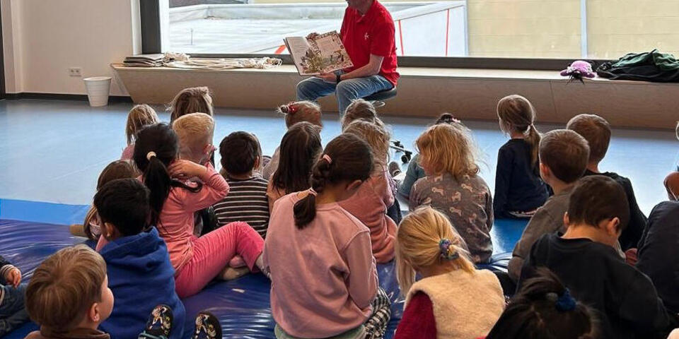 Ein Mann hält ein Buch und sitzt vor einer Gruppe von Kindern und Erwachsenen